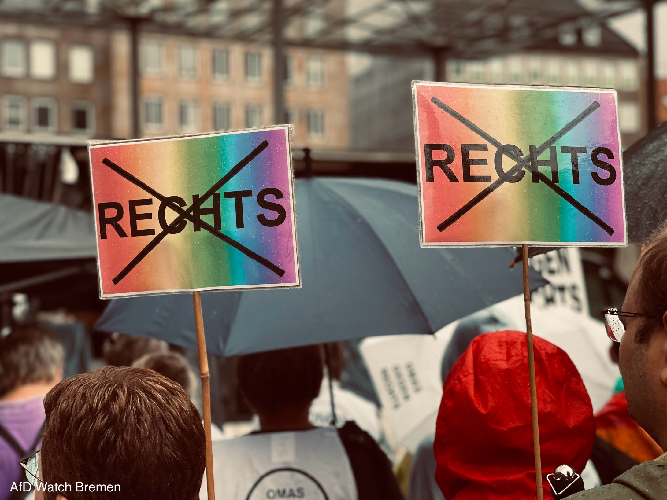 Zwei regenbogenfarbene Schilder mit dem jeweils durchgestrichenen Wort „RECHTS“ ragen über Menschen mit Regenschirmen und Regenkleidung.