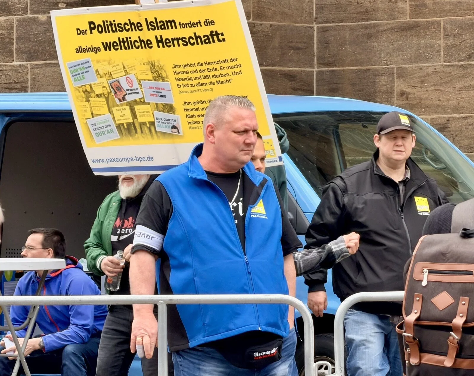 Ein Teilnehmer mit Ordner-Armbinde und blauer Weste mit BPE-Logo trägt eine schwarze Gürteltasche mit dem Schriftzug Reconquista.