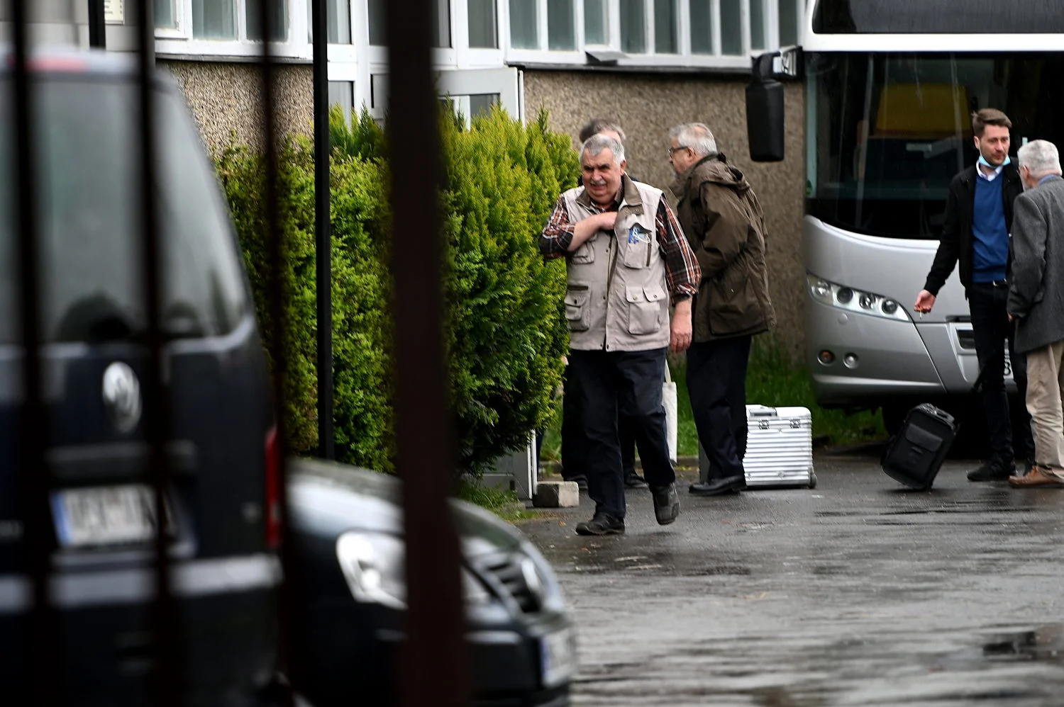 Ein Mann mit grauem Haar, beiger Weste und kariertem Hemd geht an einer Hecke entlang. Hinter ihm stehen weitere Männer neben einem Reisebus.