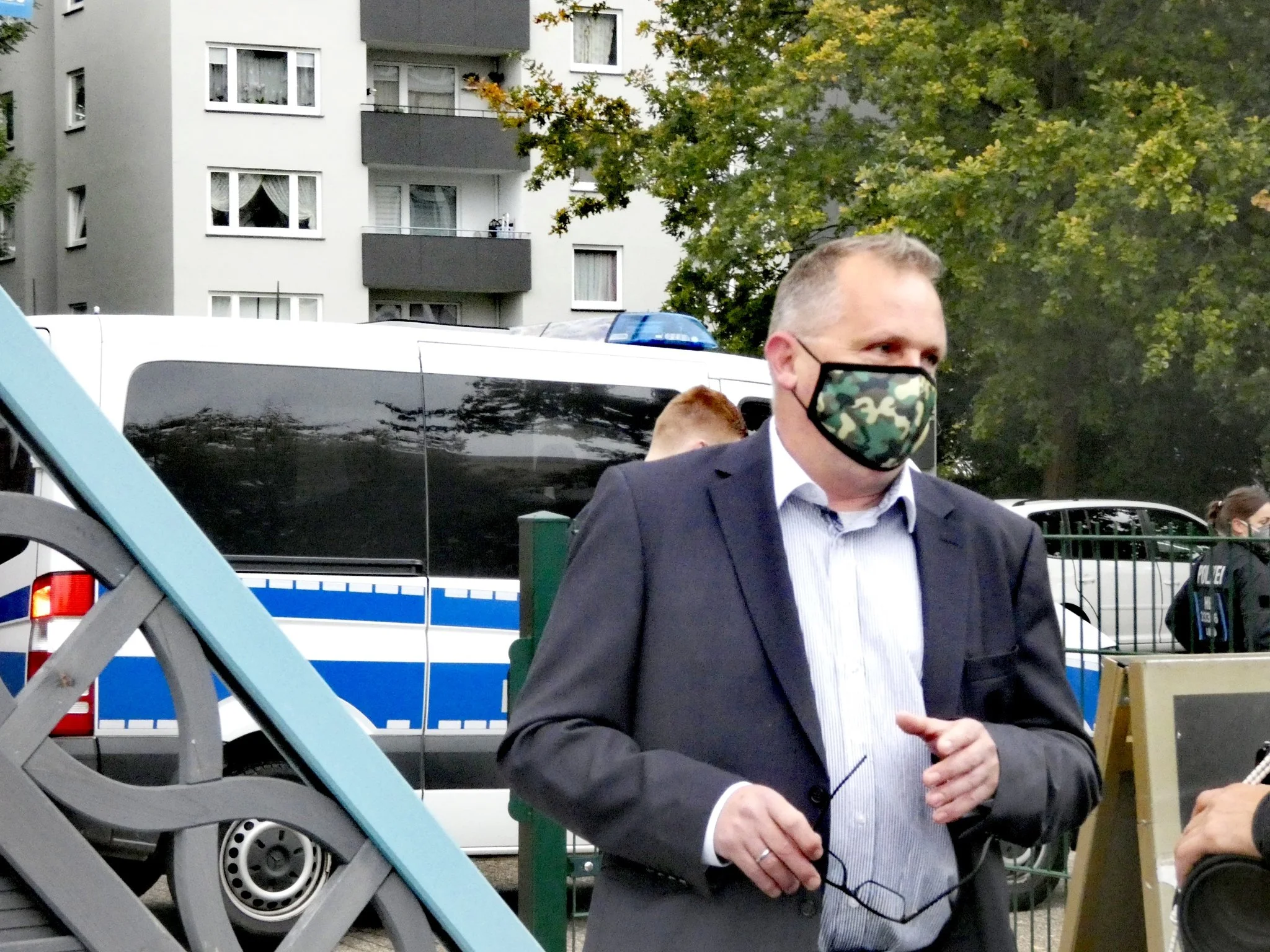 Peter Beck mit einer Maske in Tarnfarben vor dem Tagungsort; im Hintergrund steht ein Polizeifahrzeug.