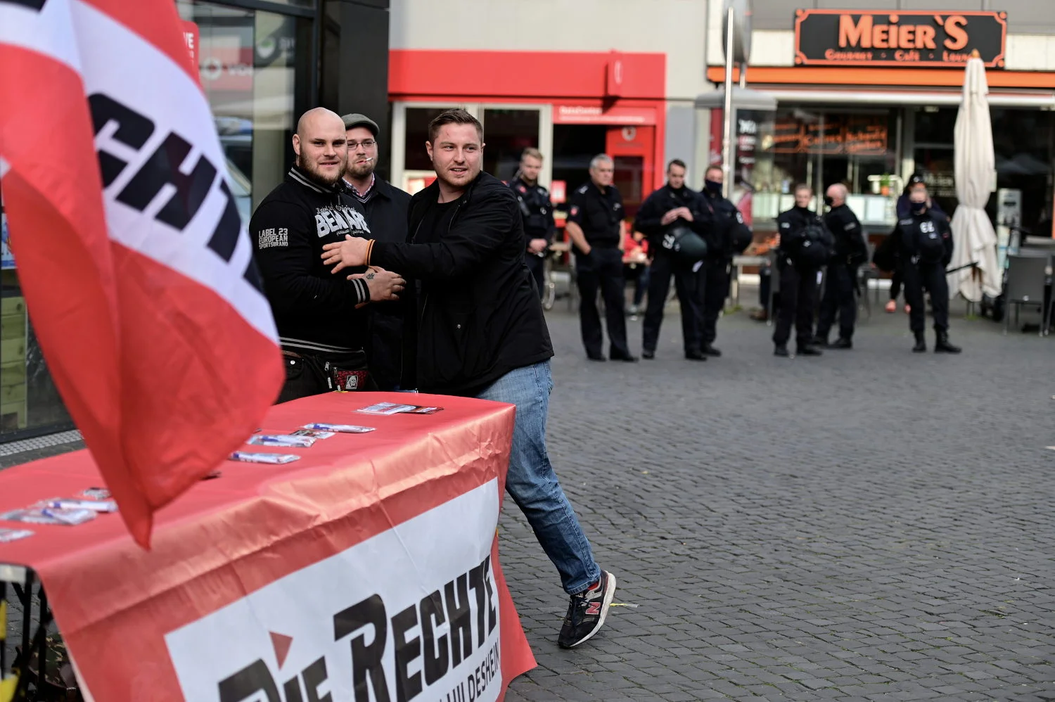 Anhänger der Partei „Die Rechte“ an einem Infostand mit Parteifahne; im Hintergrund stehen Polizeibeamt:innen.