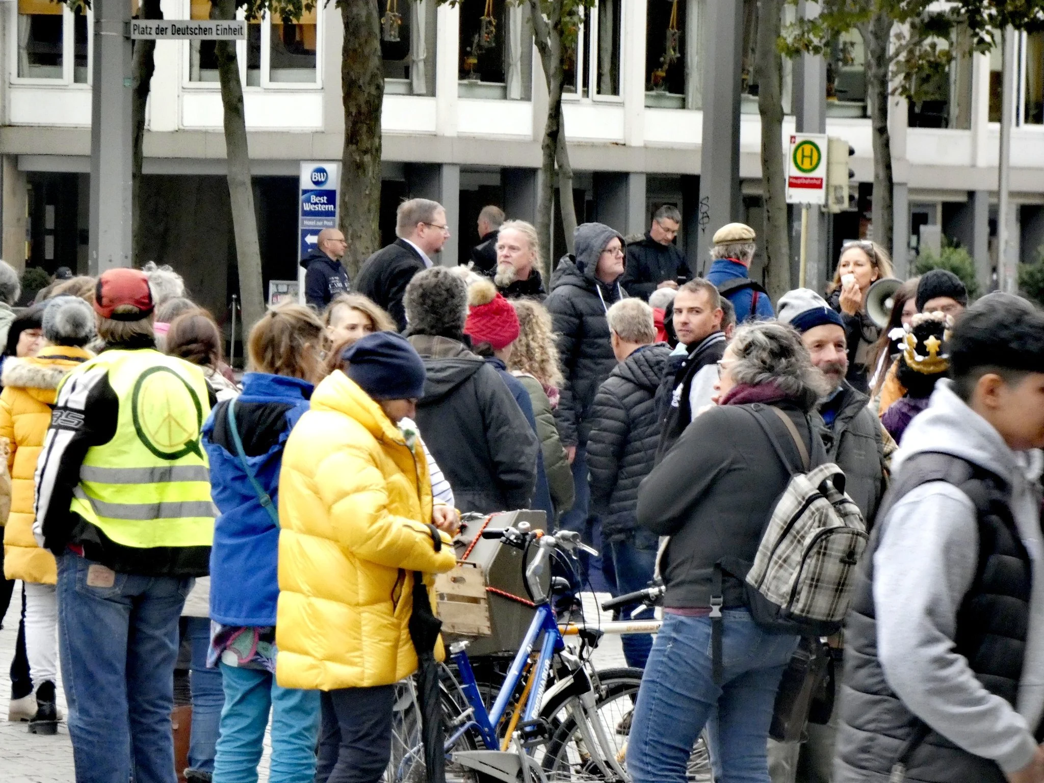 Menschen auf dem Bahnhofsvorplatz. Links trägt eine Person eine gelbe Warnweste mit einem großen Q, in das ein Peace-Zeichen eingezeichnet ist.