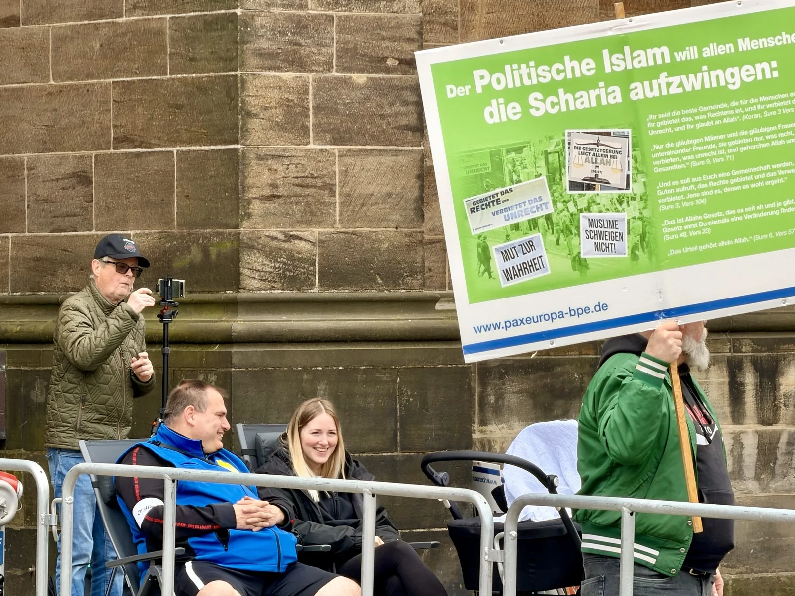 Pax-Europa-Kundgebung vor der Dommauer: Teilnehmer:innen hinter Absperrgittern und ein großformatiges Plakat mit einer Warnung vor der Scharia.