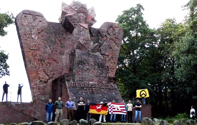 Gruppenaufnahme vor dem Niedersachsenstein: Teilnehmende halten Flaggen, darunter eine gelbe Fahne mit dem Lambda-Zeichen der Identitären Bewegung.