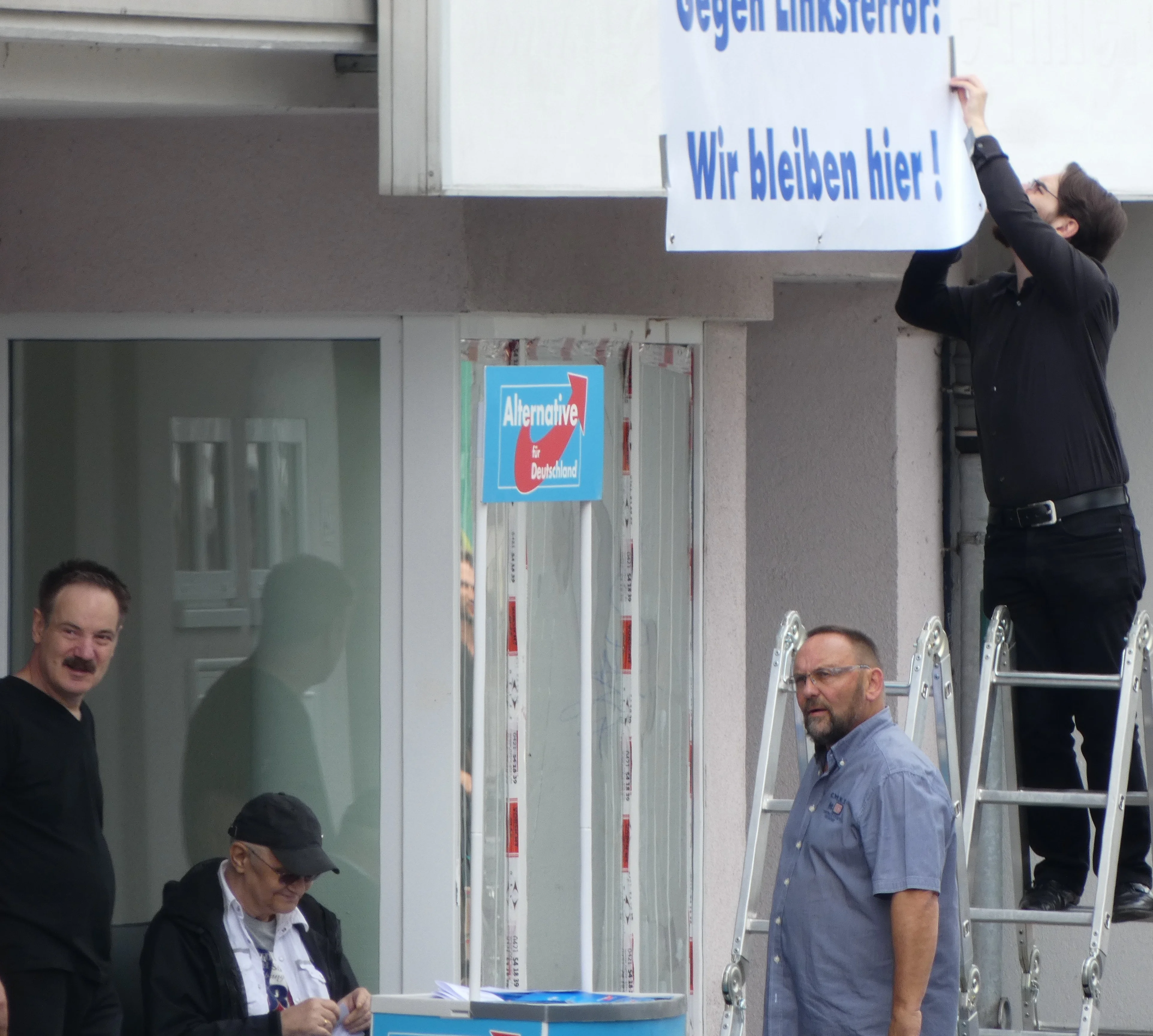 Vor der Geschäftsstelle wird ein Banner angebracht: „Gegen Linksterror! Wir bleiben hier!“. Die Bildunterschrift benennt die dokumentierten AfD-Akteur:innen.